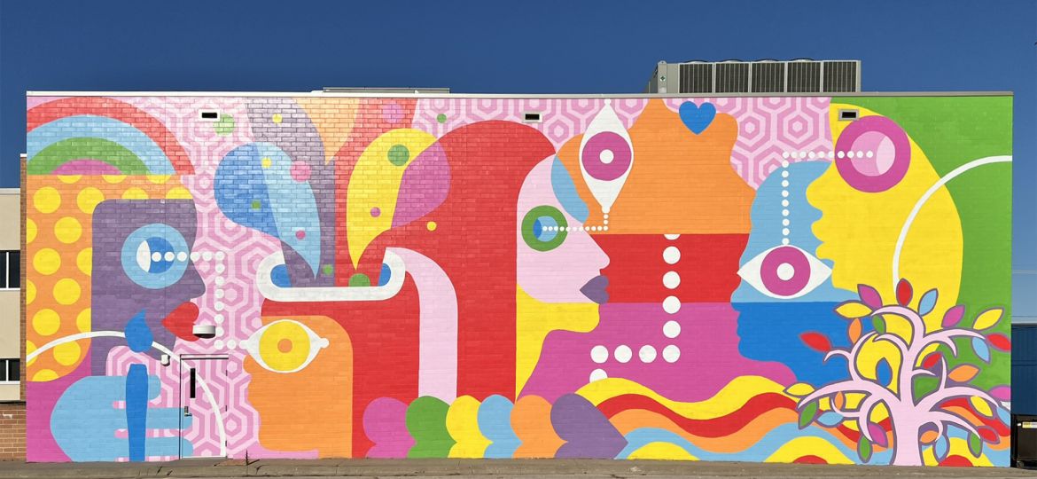 An overal view of Perspective, a colorful mural on the side of the Orpheum with geometric faces, eyes, and a stylized tree in bright pinks, oranges, yellows, and greens.