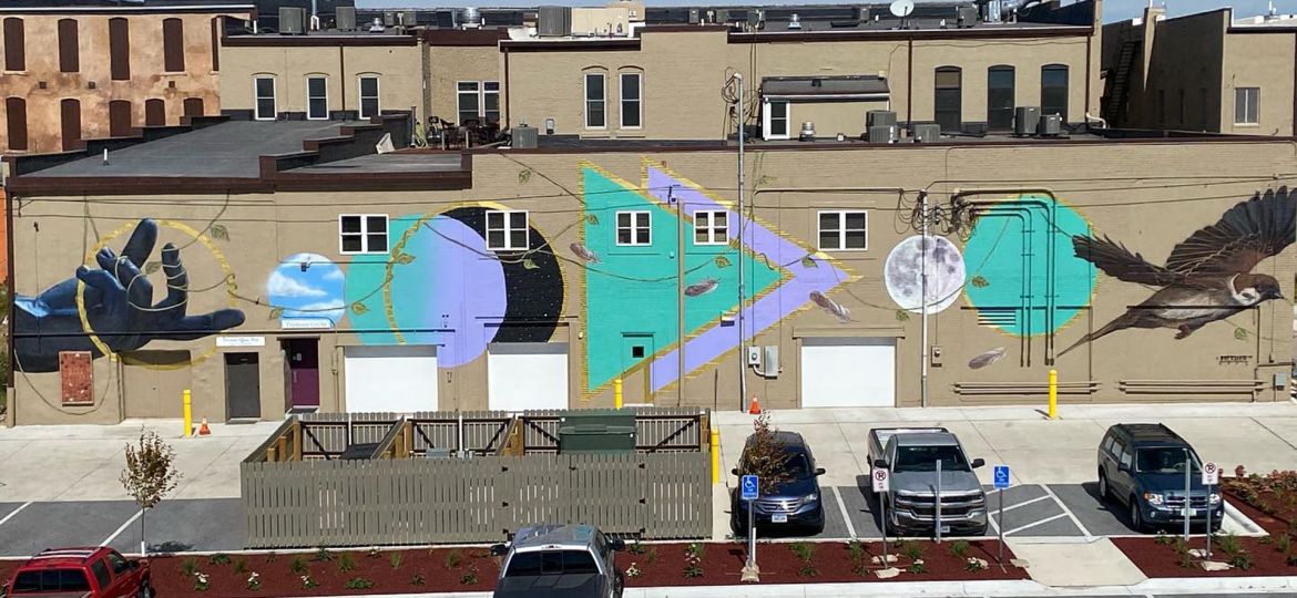 A birds eye view of the Tremont mural: far left a black hand with a vine tangled through the fingers, followed by a series of triangles and circles in purple and aqua outlined in gold, and ending with a flying bird