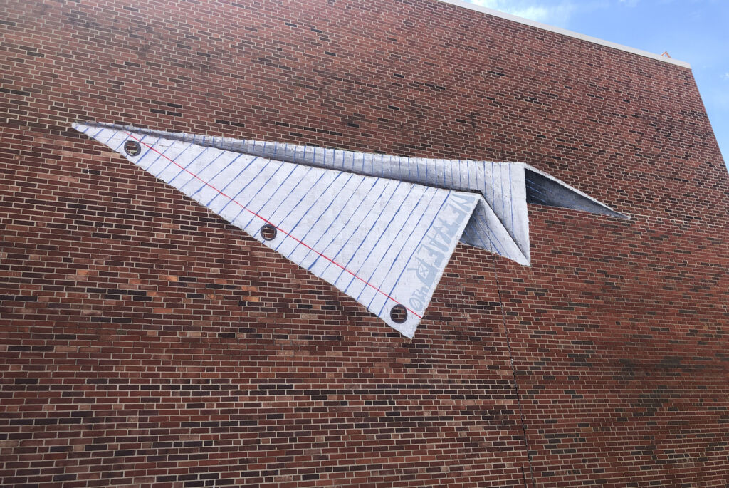 A mural of a paper airplane made from three-hole-punched lined paper on the side of a brick building