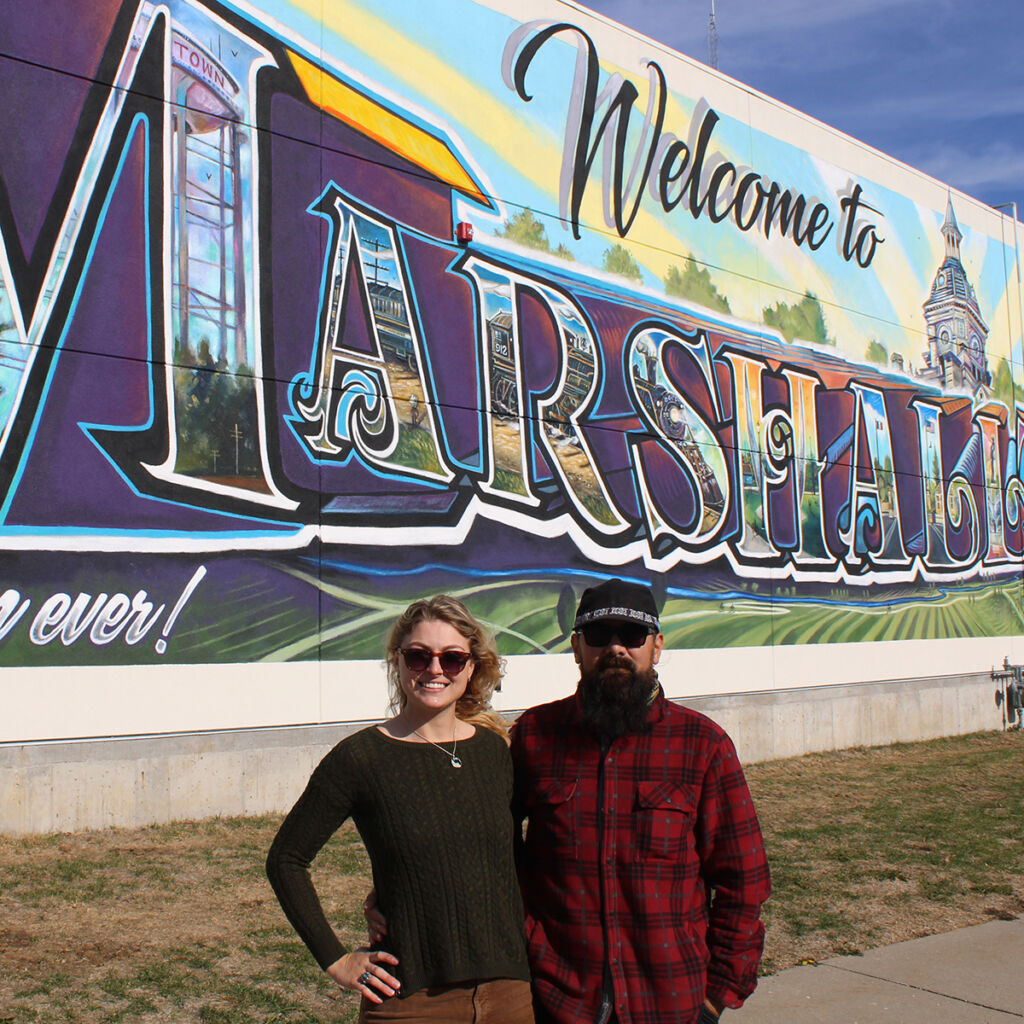 Jimmy Navarro and Katie Jenson standing in front of the Welcome to Marshalltowan postcard mural