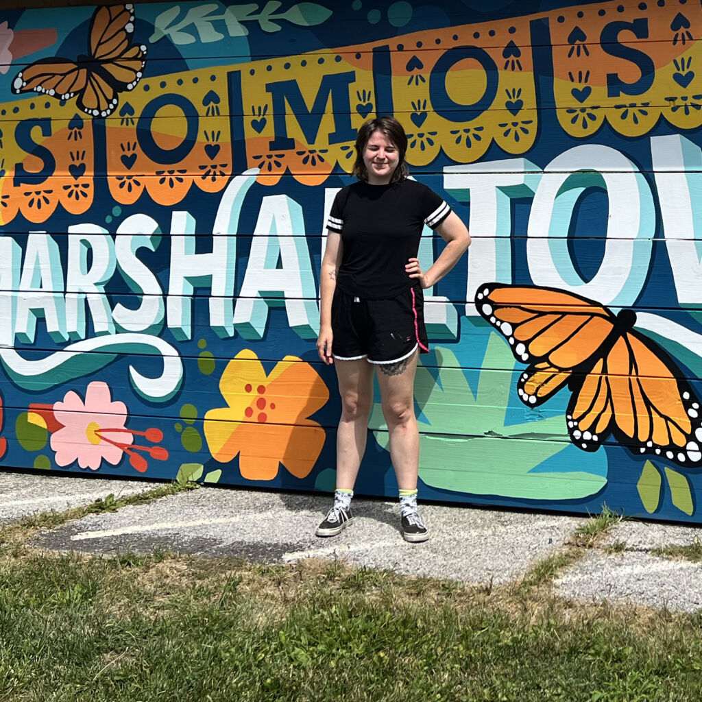 Ally stands in front of the SOMOS mural smiling with her eyes shut