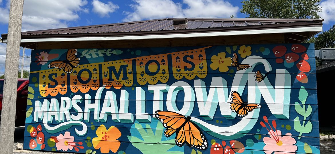 Somos Marshalltown mural with a blue backdrop and SOMOS lettering in yellow and orange flag style lettering with Marshalltown in white lettering below. Monarch butterflies and stylized flowers surround the lettering